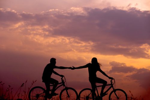 Two people riding bicycles in single file against sunset sky reaching out to one another