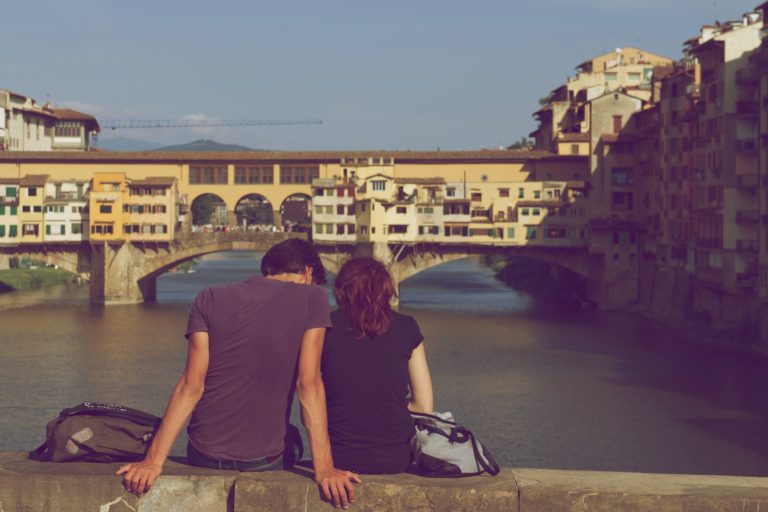 Young couple sitting on a bridge looking towards a river and buildings in the background.