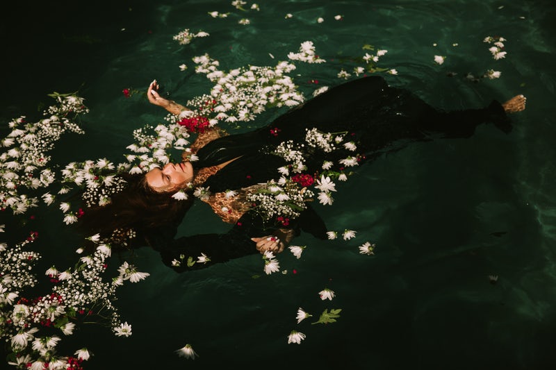 A woman in a black dress with her arms outstretched and her face up floats in green water surrounded by white and red flowers.
