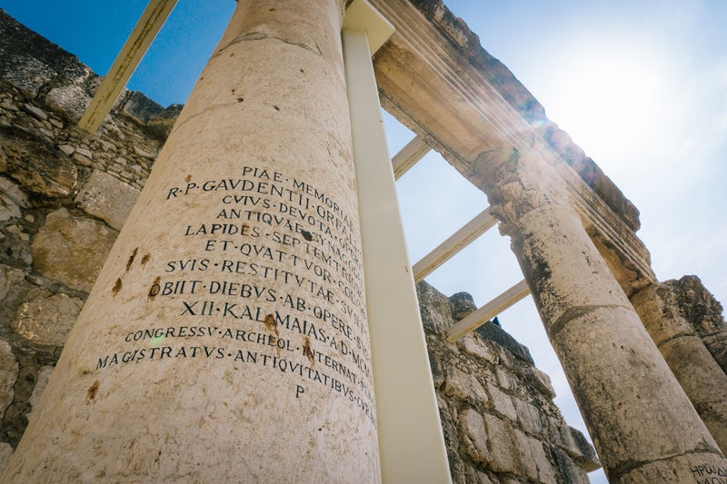 Greek words are etched into an Ancient Greek column and portico in front of sunlight and sky.