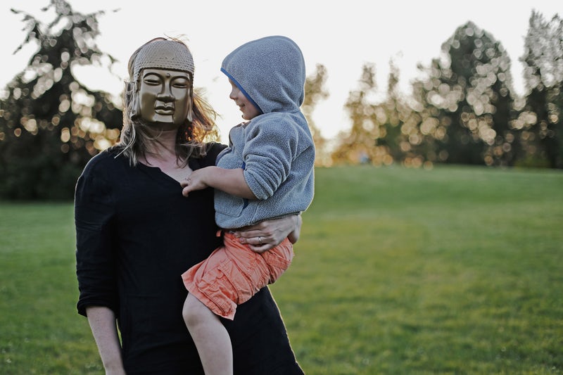 A boy in a hoodie smiling as he looks at the face of a woman holding him, who is wearing a golden buddha face mask