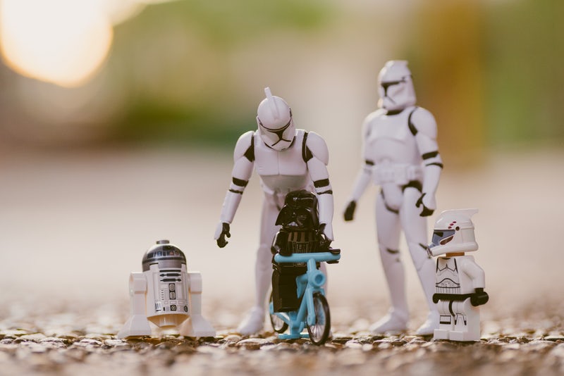 Two larger Star Wars figurines positioned above smaller Star Wars figurines, one of them riding a blue tricycle