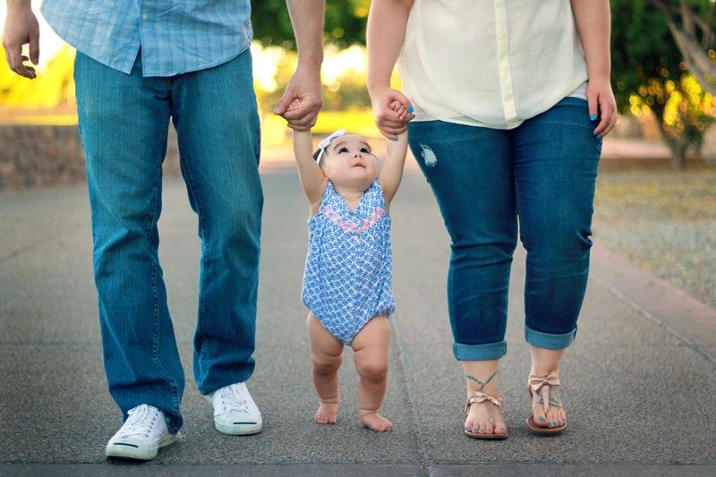 Two adults from the waist down walking, each holding one of the hands of a toddler in a blue onesie who is barefoot and walking as she looks up at them