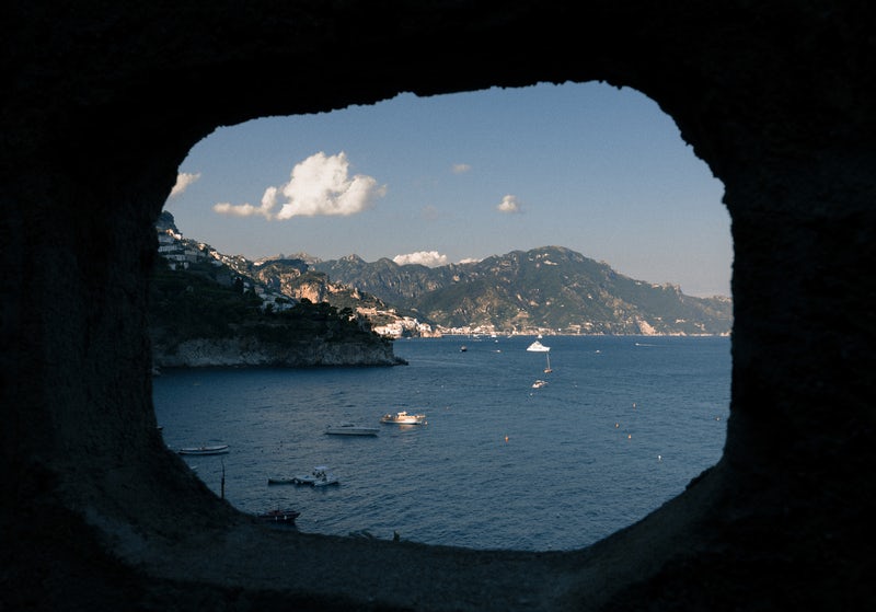 Through a circular cave's opening, white clouds float above mountains and boats in a harbor.