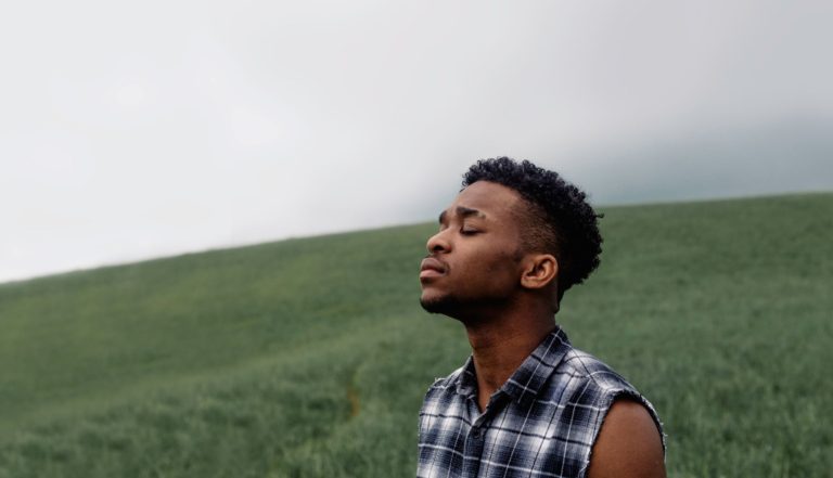 A black man stands in a field with his eyes closed. His head is tilted towards a cloudy sky.