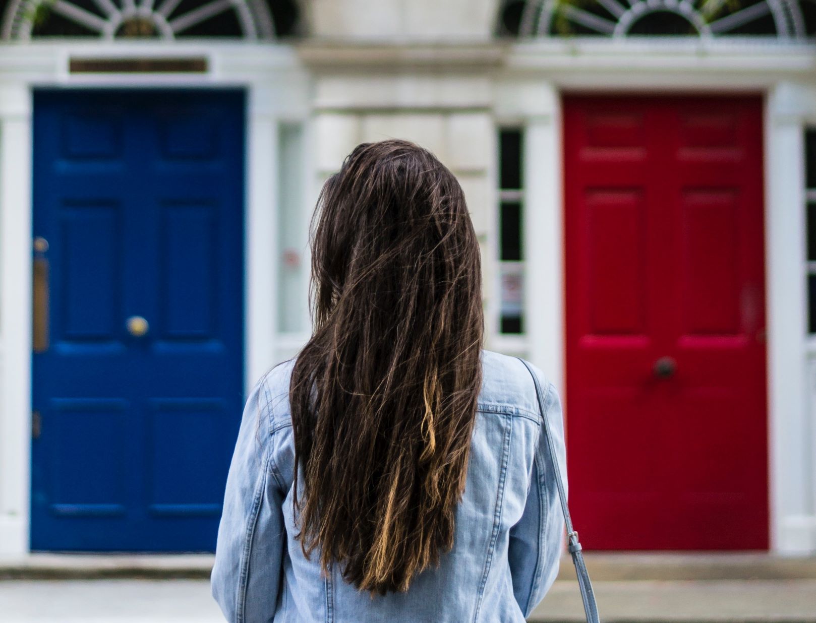 Woman with dark brown hair has her back to the camera. She's wearing a jean jacket trying to decide if she should go through the blue door on the left or the red door on the right.