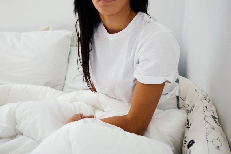 An unsmiling woman from the chin down sitting in a white bed wearing a white t-shirt.
