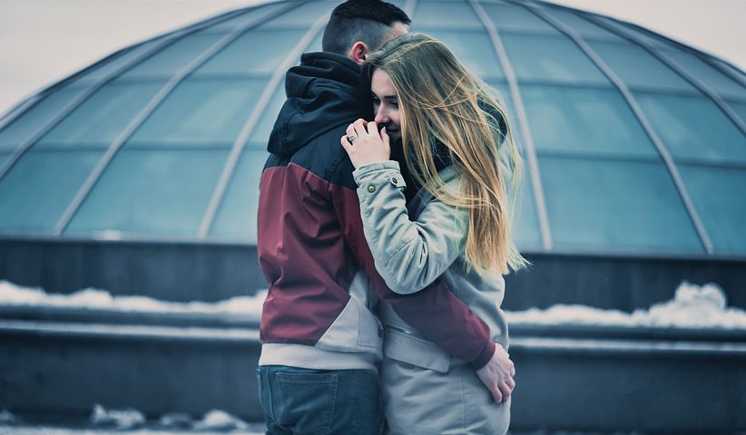 Young man and woman embracing, facing different directions in front of snow covered roof.